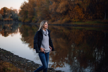 Fototapeta na wymiar Adorable blonde woman walking along river looking away smiling with black jacket on shoulders, hands on sweater with trees covered with sunset in background. Autumn walk in forest.