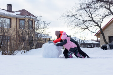 Little girl with mother making massive snow ball for building snowman on backyard in evening with trees and house in background. Parents spending time with children.