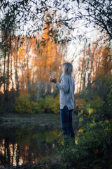 Peaceful blonde woman standing by river in forest, looking away at sunset and posing with trees in background being part of nature, wearing blue jeans and grey sweater. Autumn walk in forest.