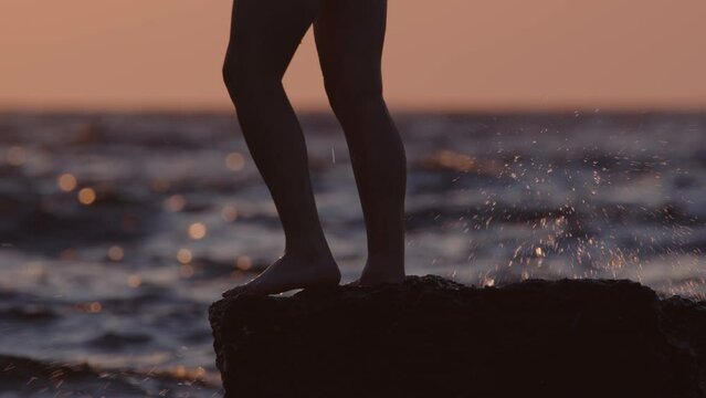 Man dives into water from rock to swim in sea at sunset. Close-up of male legs standing on edge of stone reef jumping into ocean.