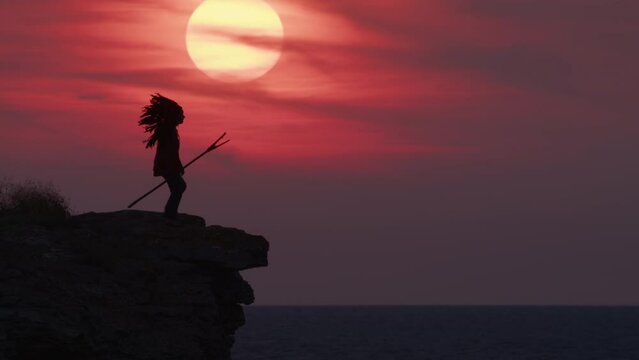 Silhouette of Red Indian chief in headdress with feathers approaches edge of cliff on shore of ocean or sea and looks into distance on background of amazing sunset sky with sun.