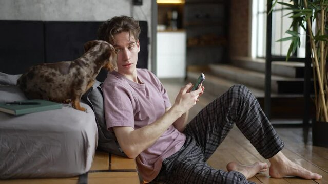 4k Young man is using smartphone and having good time with dog in apartment spbd. Closeup view of caucasian guy holds phone in his hand and looks with smile, talks with cute pet and sits in light