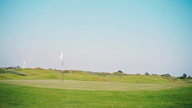 Static meditative footage. A white flag on a golf hole flutters in the wind. Green bright grass on the golf course against the blue sky