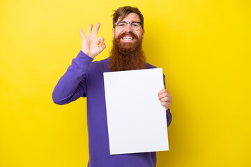 Redhead man with beard isolated on yellow background holding an empty placard and doing OK sign