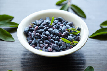 Haskap berry with fresh green leaves in white bowl on wooden table. Honeyberry (Lonicera caerulea)