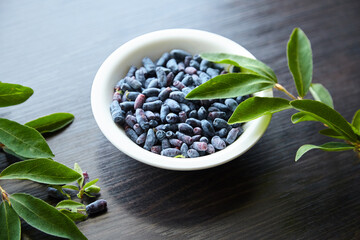 Haskap berry with fresh green leaves in white bowl on wooden table. Honeyberry (Lonicera caerulea)