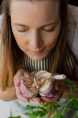 Vertical closeup woman face with close eyes holding and smelling garlic, greenery on table. Healing...