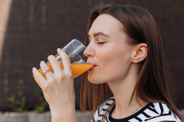 Portrait of beautiful young woman drinking and taking sips of fresh cold orange juice outdoor on terrace closeup side view. Lady with closed eyes enjoying taste of drink, refreshing on summer day