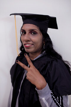 Young woman wearing graduation cap and ceremony robe smiling and confident gesturing with hand doing victory sign with fingers looking and the camera