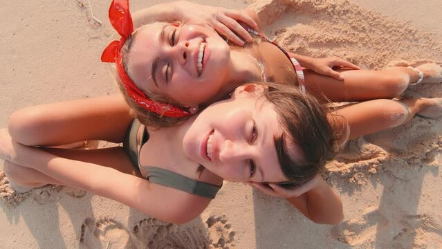 Young attractive Caucasian women sunbathing sits on sand and leaning back on each other's backs and looking up dressed in beach swimsuits spending holidays on sunny sea coast or resort. Top view