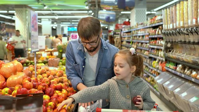 Father and little girl in shopping cart choose red apples in vegetable section in supermarket. Shopping products in store. Buying food, fruits