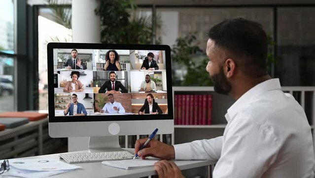 Video call, online conference. Over shoulder view of Indian man, on computer screen, talks with multinational group of successful business people, virtual business meeting, telecommunication concept