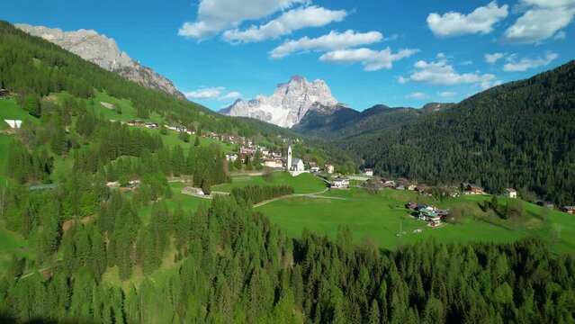 AERIAL: Beautiful view of mountain village with high alpine peaks in background. Picturesque sight of alpine village surrounded with woodland and high snow-capped mountains in springtime on sunny day.