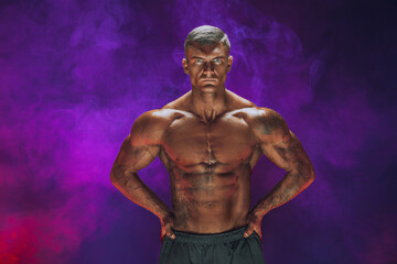 Fototapeta na wymiar A handsome confident athletic man with bare mscular torso posing on a dark background filled with smoke. Male beauty concept. Fitness, bodybuilding