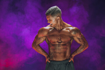 Fototapeta na wymiar A handsome confident athletic man with bare mscular torso posing on a dark background filled with smoke. Male beauty concept. Fitness, bodybuilding