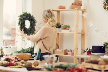 Blonde young woman in sweater standing in own workshop and looking at green Christmas wreath