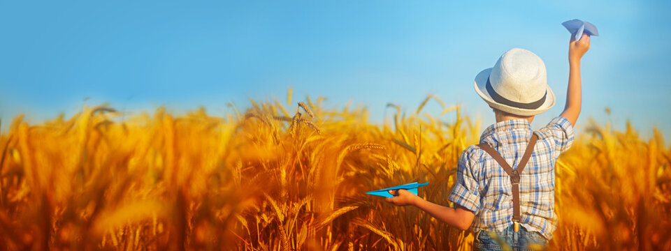 Happy child with a toy paper airplane on a sunset background over a wheat field. Back view. Childhood concept. Banner.