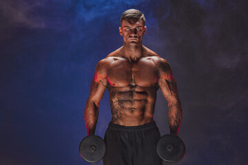 Fototapeta na wymiar Brutal sweaty strong young man athlete with naked upper body standing doing workout with dumbbels and showing strong pumped up biceps over smoky background. Sport men body concept