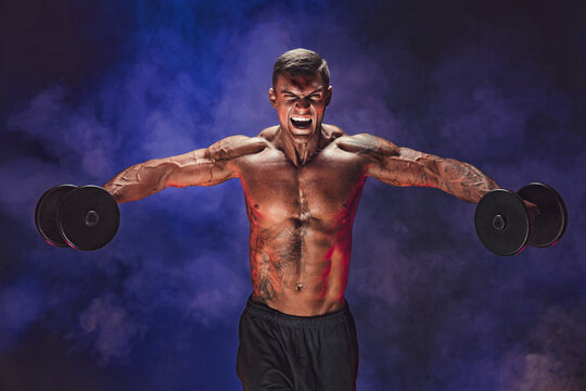 Brutal sweaty strong young man athlete with naked upper body standing doing workout with dumbbels and showing strong pumped up biceps over smoky background. Sport men body concept