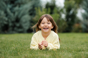 Funny smiling blonde little child girl 4-5 year old lying on green grass lawn outdoor. Small kid...