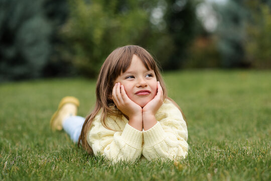 Funny smiling blonde little child girl 4-5 year old lying on green grass lawn outdoor. Small kid wear knitted casual sweater over nature background in park. Childhood. Autumn season.
