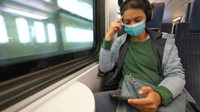 Commuter woman riding train looking at smartphone device wearing covid face mask and headphones