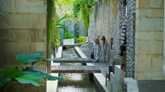 backs of European ladies walking in white long dresses down a narrow street with a small river with green plants