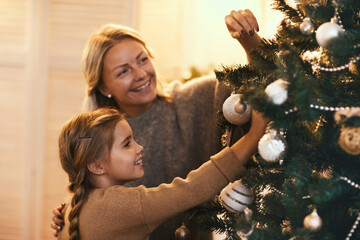 Happy beautiful mother and daughter decorating coniferous tree with baubles while preparing home...