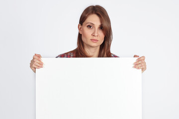Young attractive woman with dark brown hair and eyes holds empty white board or banner with great copy space for any text or slogan. Serious look, confident face expression, concernity concept.