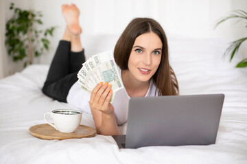 Young girl lying on a bed and holding polish zloty, uses laptop computer. Work from home