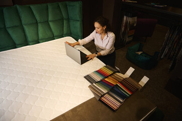 Top view of inspired interior designer working on laptop on orthopedic mattress near spread out various colorful fabric swatches in home interior design studio