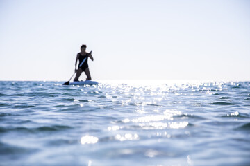 Silhouette of a woman on sap board with a paddle in her hands against a background of blue water and sky. The concept of conquering the elements. The idea of an active lifestyle. Horizontal photo with