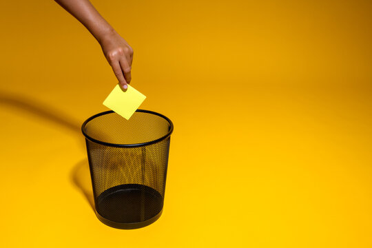 The hand throws a yellow sticker into the trash. A piece of paper is thrown into the office wastebasket. The concept of getting rid of the use of paper in the office