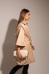 beautiful woman in brown coat, black pants, white boots holding a white bag posing to the camera