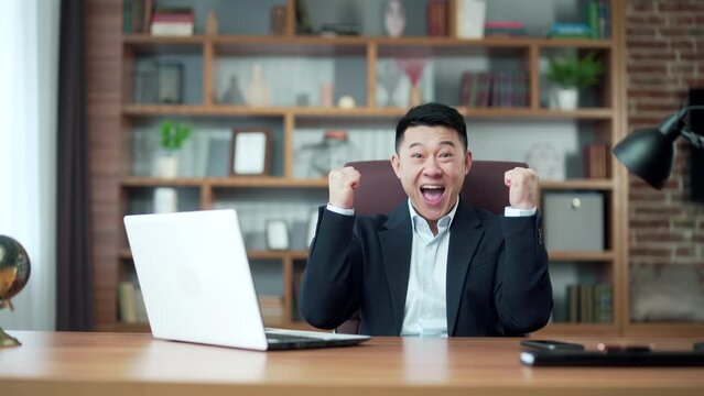 excitable asian man screams with joy and waves hands looks at laptop screen after winning online casino and getting jackpot sits in office. Gambling applauds good news result. looking at camera
