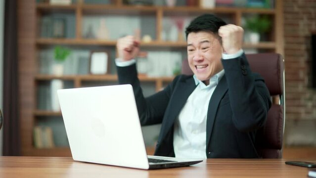 Young excitable asian man screams with joy and waves hands looks at laptop screen after winning online casino and getting jackpot sits at table in office. Gambling addiction applauds good result. Asia