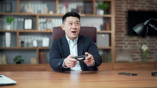 Asian man in suit in office at work playing computer games on console joystick, sitting at table desktop, smiling, happy, relaxed. Cheerful business man during a break in free time videogames