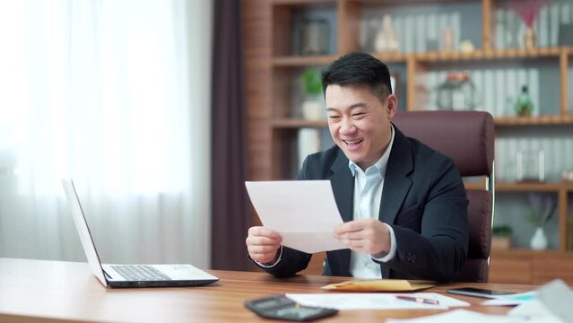 Excited satisfied asian businessman in suit reads a document. happy man looking, checking at paper letter correspondence with good news result, happy. getting bank loan approvement notification. Asia