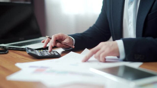 hands close up of financial analyst accountant at desk counting report on calculator with documents and graphs. busy. working Desktop at computer desk. business manager man in suit in office. indoor