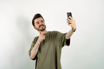 Cheerful caucasian man wearing casual clothes posing isolated over white background pointing index finger on mobile phone while taking selfie with big smiles. Communicating concepts.