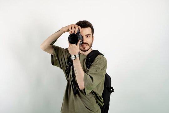 Portrait of cheerful man wearing khaki t-shirt posing isolated over white background taking images with dslr camera. Photographer covering his face with the camera.