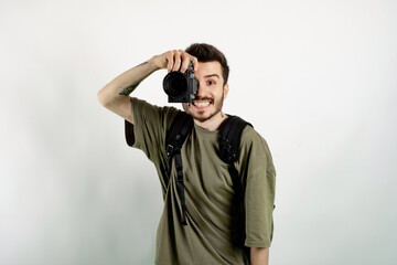 Cheerful young man wearing t-shirt posing isolated over white background taking images with dslr camera. Photographer covering his face with the camera.