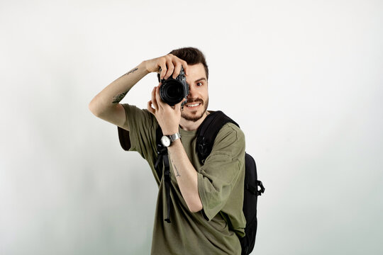 Cheerful caucasian man wearing casual clothes posing isolated over white background taking images with dslr camera. Photographer covering his face with the camera.