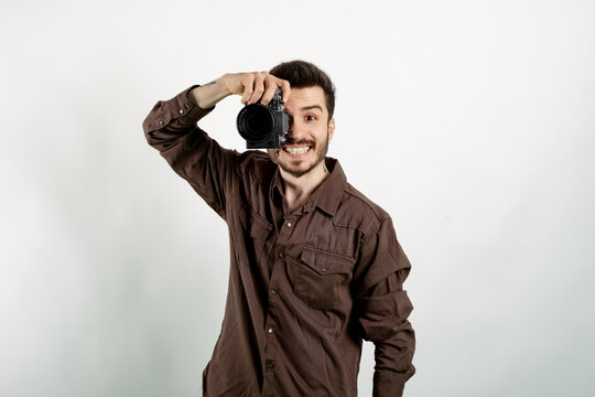 Happy casual man wearing casual clothes posing isolated over white background taking images with dslr camera. Photographer covering his face with the camera.