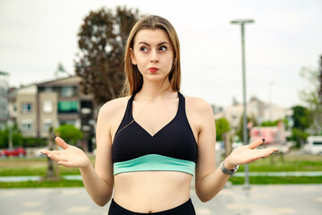 Cute brunette woman wearing sports bra standing on city park, outdoors clueless and confused with open arms, shrugging shoulders, no idea and doubtful face.