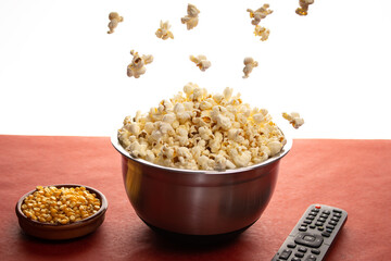 popcorn falling into an aluminium bowl, with a TV remote control on one side and an earthenware bowl with kernels of corn inside on a red table with a white background.