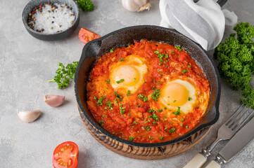 Shakshuka in a frying pan on a gray concrete background with ingredients. Eggs cooked in tomato sauce with spices and fresh parsley on top. Copy space.