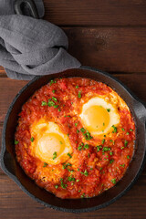 Shakshuka in a frying pan on a dark wooden background. Eggs cooked in tomato sauce with spices and fresh parsley on top. Rustic style. Top view, copy space.