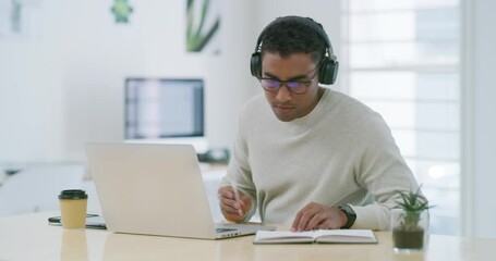 Mixed race creative entrepreneur using a laptop, writing notes in a notebook while listening to music on headphones. Young latino businessman and designer sitting alone and working from home office - Powered by Adobe
