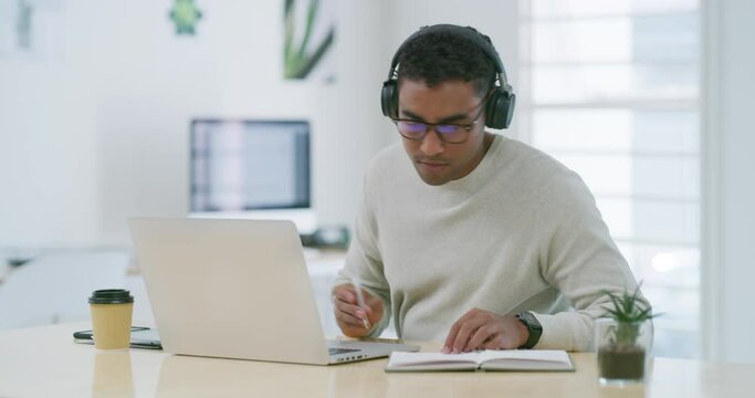 Mixed race creative entrepreneur using a laptop, writing notes in a notebook while listening to music on headphones. Young latino businessman and designer sitting alone and working from home office